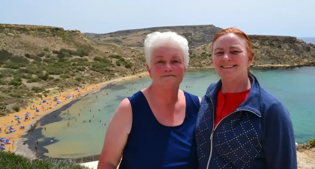 Two people smiling at Ramla Bay beach, Malta, with a scenic view of the sandy shore and turquoise water in the background.