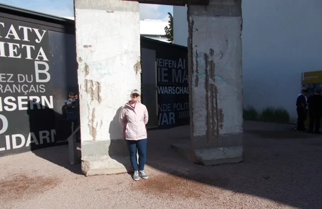 Person standing by Berlin Wall section, outdoor exhibit with text and shadow.