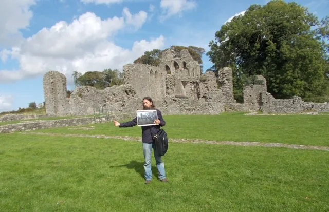 Tourist holding a photo in front of scenic ancient ruins on a sunny day with blue sky and lush greenery.