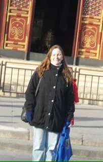 Person standing in front of a traditional ornate building facade, wearing a black coat and holding a blue bag.
