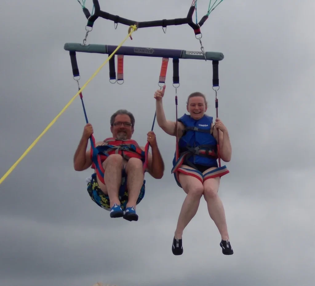 Two people parasailing with smiles in the sky, wearing blue life vests and harnesses on a cloudy day.