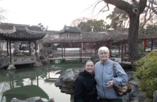 Two people smiling at a traditional Chinese garden with a pond and pavilion in the background.
