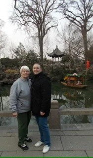 Two people standing in front of a serene park pond with a traditional gazebo and trees in the background.