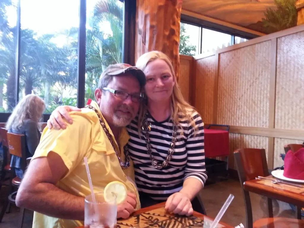 Smiling couple sitting at a restaurant table with drinks, wearing leis, tropical backdrop visible through window.