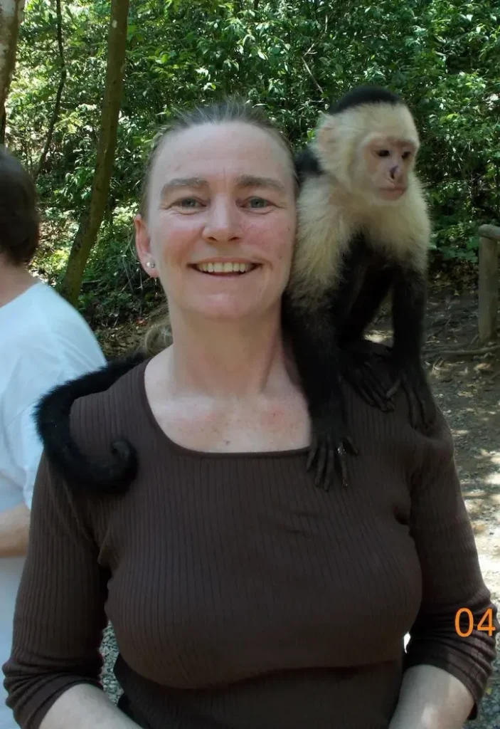 Woman smiling with a capuchin monkey on her shoulder, standing amid lush green trees.