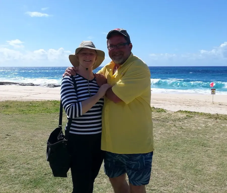 A smiling couple embraces on a sunny beach, with waves in the background and clear blue skies overhead.