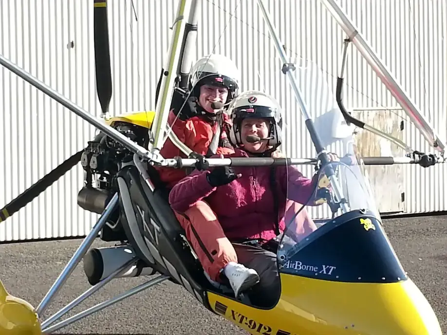 Two people in helmets ready for a microlight flight, smiling and seated in a light aircraft by a hangar.