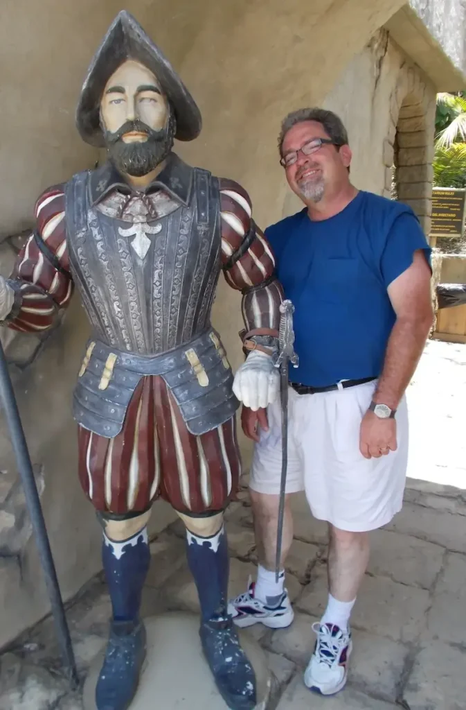 Man posing with historic explorer statue, wearing a blue shirt and white shorts.