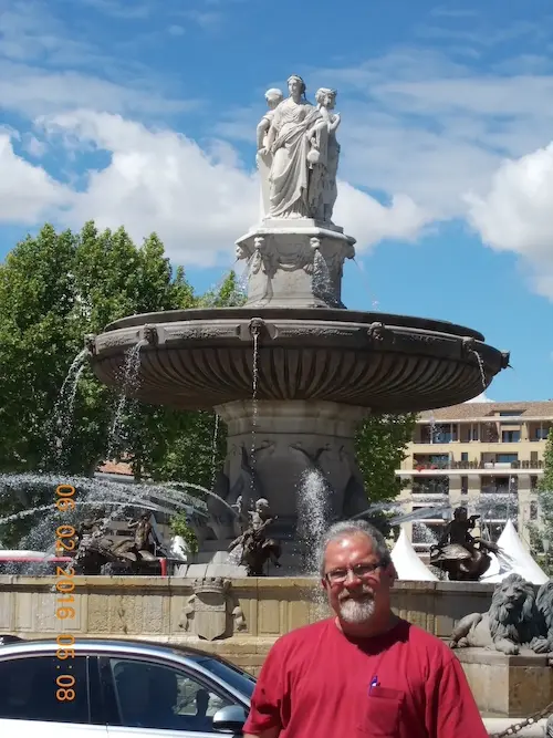 Man smiling in front of a historic fountain with sculptures, set against a bright blue sky and trees.