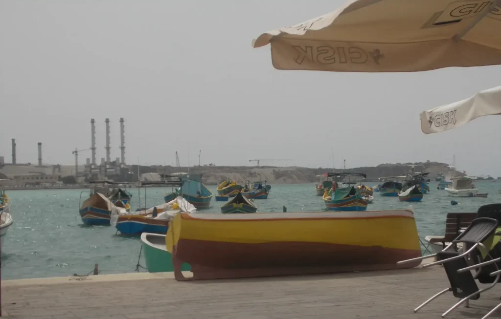 Colorful boats float in a harbor under a clear sky, with industrial buildings in the background.