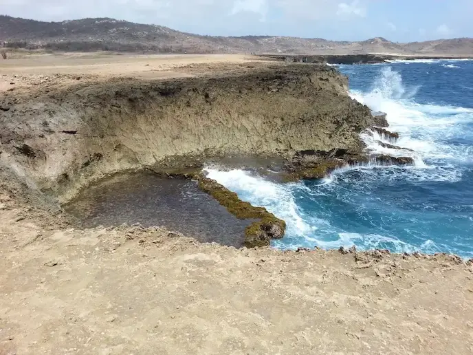 Rocky coastal cliff with waves crashing against the shore and a natural pool formation.