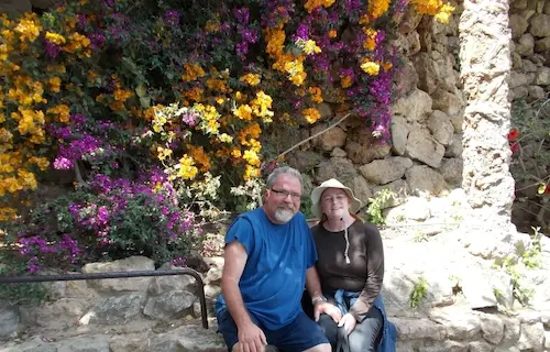 Couple sitting by a stone wall with vibrant purple and yellow flowers in a sunny garden setting.