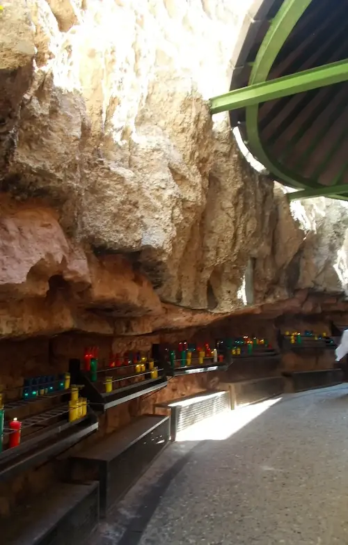 Candles placed on shelves carved into a rocky wall, brightly lit space with sunlight filtering through a green overhang.