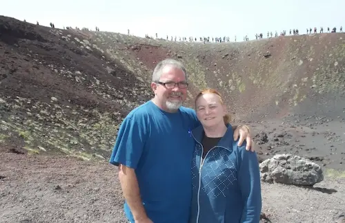 Couple smiling at a volcanic crater, surrounded by rugged terrain and people in the background under a clear sky.