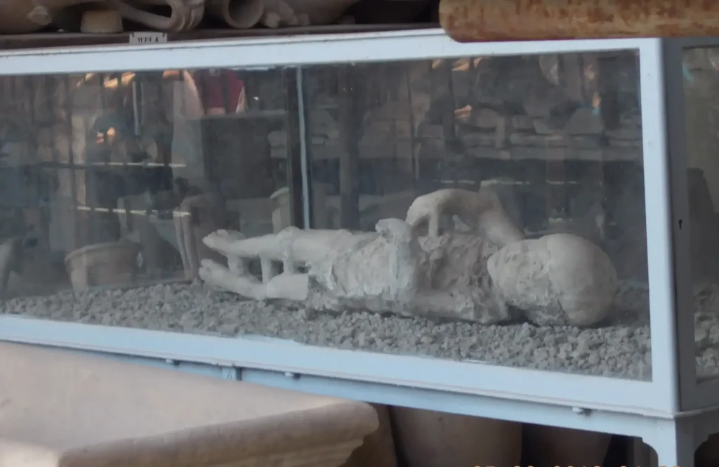 Plaster cast of a victim from Pompeii eruption displayed in a glass case, surrounded by ancient artifacts.