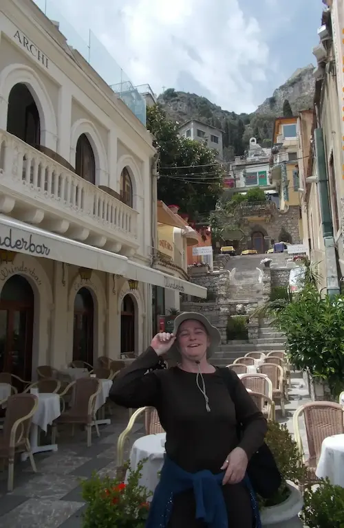 Woman posing in front of a charming café on a European street with colorful buildings and stairs in the background.