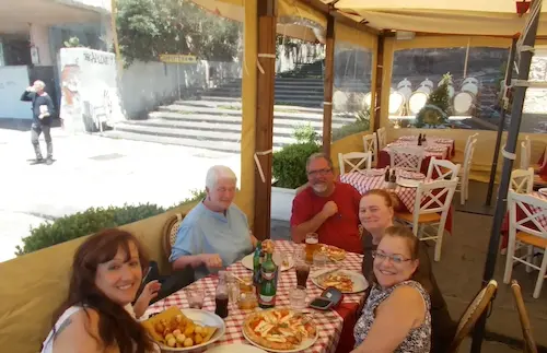 A group of people enjoy a meal together at an outdoor restaurant with red checkered tablecloths on a sunny day.
