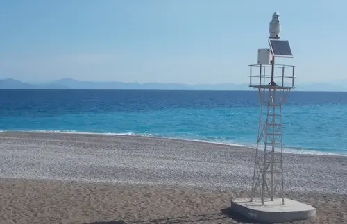 Beach with clear blue sea and small lighthouse structure under a sunny sky, offering a peaceful coastal view.